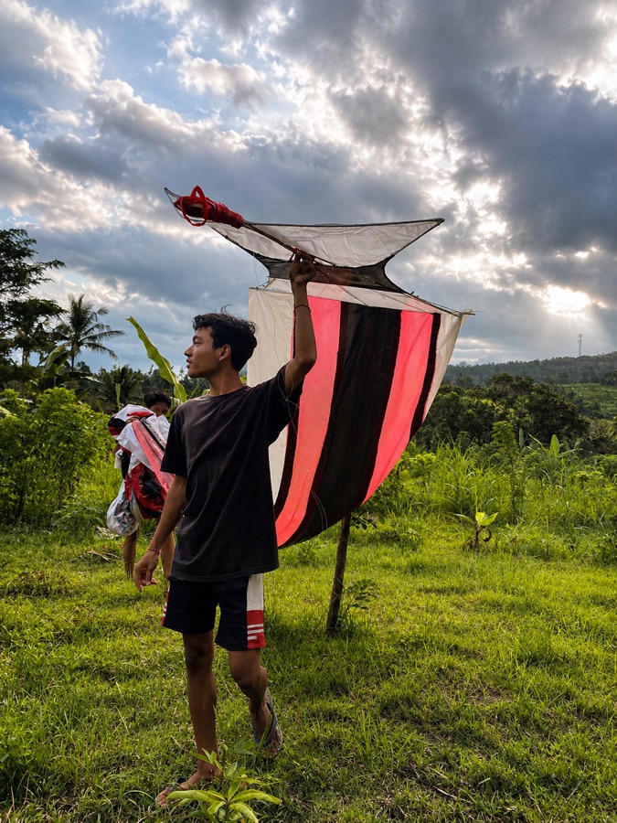 Young person carrying a large handmade kite through green fields in Karangasem
